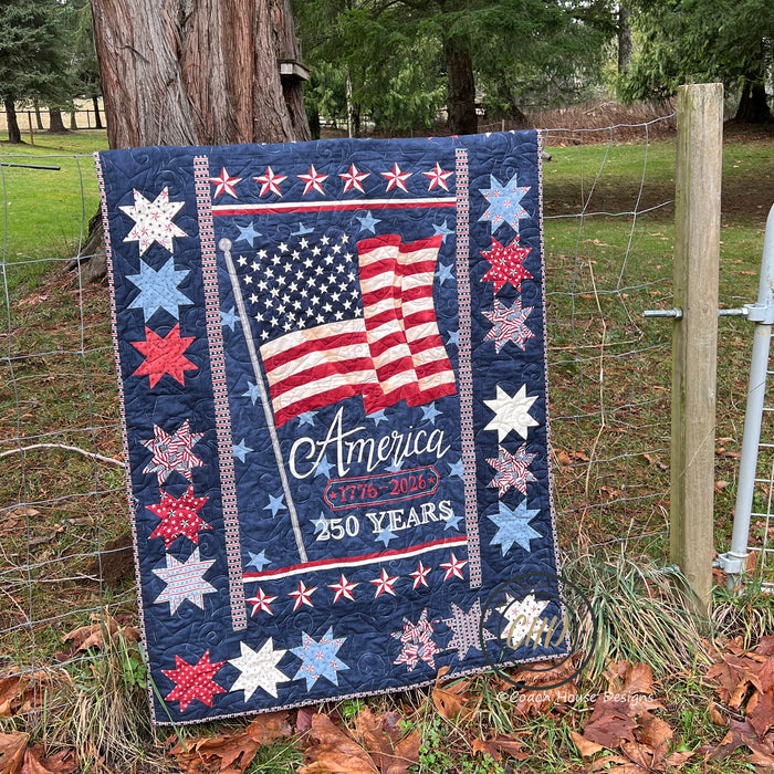 A patriotic lap quilt hangs on a country fence. The quilt is titled Celebrating 250 by Coach House Designs. The central printed panel has a proud American flag, blowing in the wind. The text states America 1776-2026, 250 years.  This is the darker panel option of this quilt pattern, with the flag and text on a blue background. It is  surrounded by a wide blue border with patriotic stars in red white and blue.