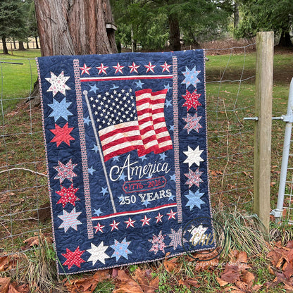 A patriotic lap quilt hangs on a country fence. The quilt is titled Celebrating 250 by Coach House Designs. The central printed panel has a proud American flag, blowing in the wind. The text states America 1776-2026, 250 years.  This is the darker panel option of this quilt pattern, with the flag and text on a blue background. It is  surrounded by a wide blue border with patriotic stars in red white and blue.