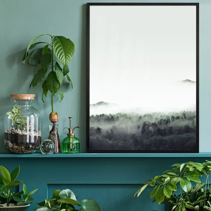 Misty forest print with subtle green tones in a black frame, displayed on a teal shelf with plants.