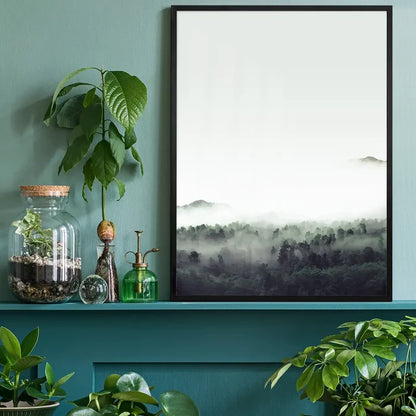 Misty forest print with subtle green tones in a black frame, displayed on a teal shelf with plants.