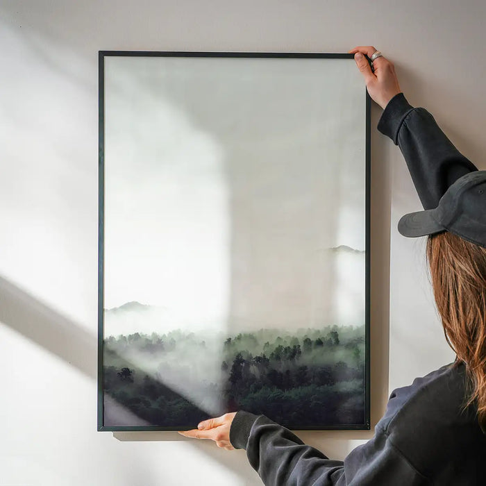 Framed photographic forest print with subtle green tones being hung on a white wall.