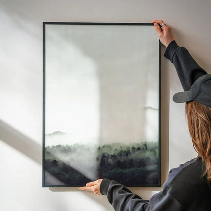 Framed photographic forest print with subtle green tones being hung on a white wall.