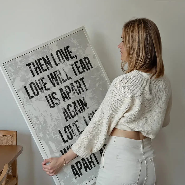 Woman holding a framed white grunge art print with bold black "Love Will Tear Us Apart" lyrics.