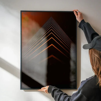 Person hanging a framed orange pyramid geometric print on a white wall.