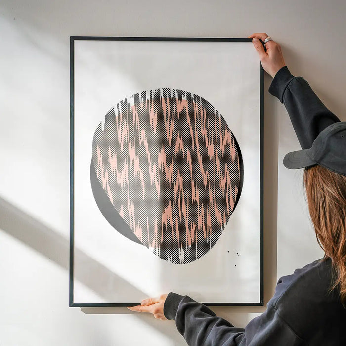 Person hanging a framed Pebble 1 abstract print with a black circle and pink patterned fill on a white wall.