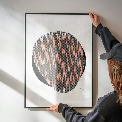 Person hanging a framed Pebble 1 abstract print with a black circle and pink patterned fill on a white wall.