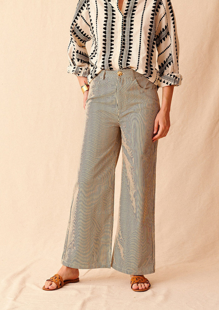 A woman standing wearing cream and black pinstriped wide leg jeans with brown sandals and cream and black embroidered top