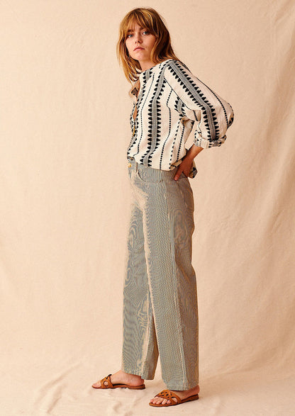 A woman standing wearing cream and black pinstriped wide leg jeans with brown sandals and cream and black embroidered top, seen from side