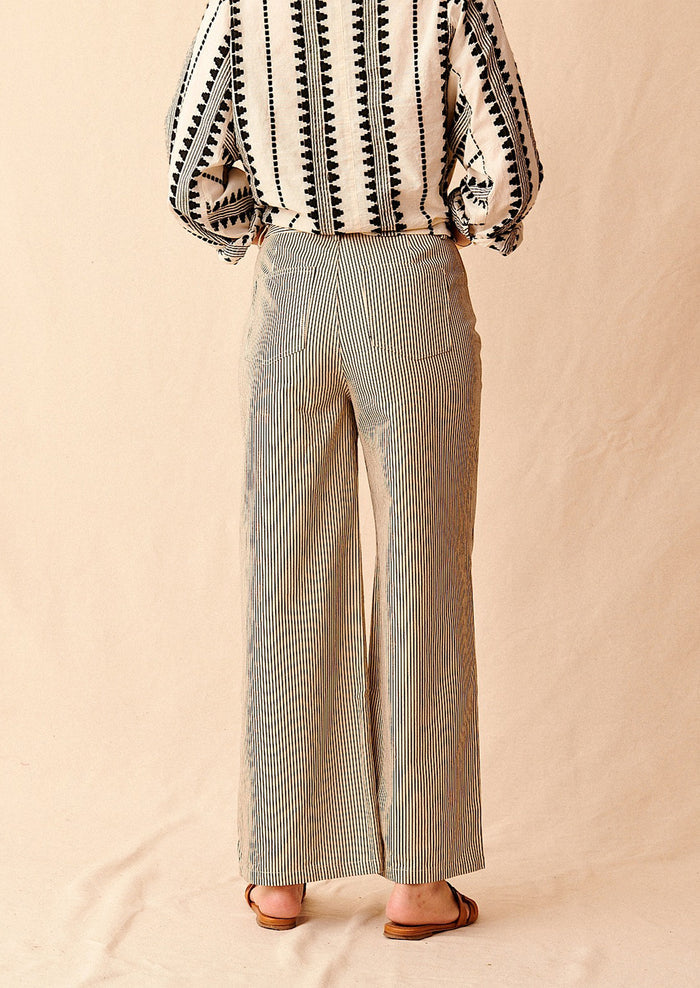 A woman standing wearing cream and black pinstriped wide leg jeans with brown sandals and cream and black embroidered top, seen from behind