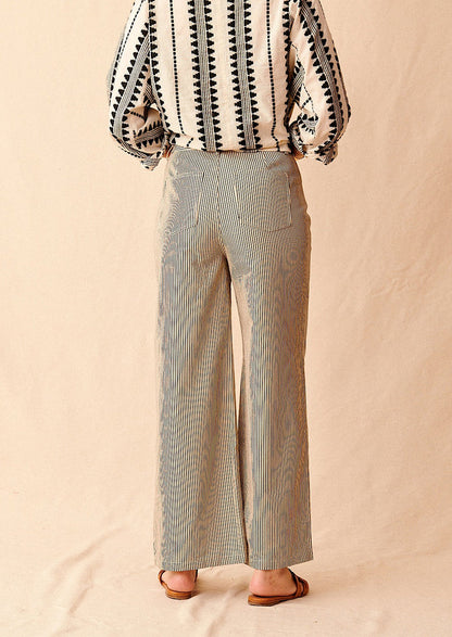 A woman standing wearing cream and black pinstriped wide leg jeans with brown sandals and cream and black embroidered top, seen from behind