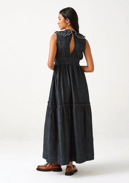 A woman standing wearing a long denim dress with an embroidered collar, viewed from back.