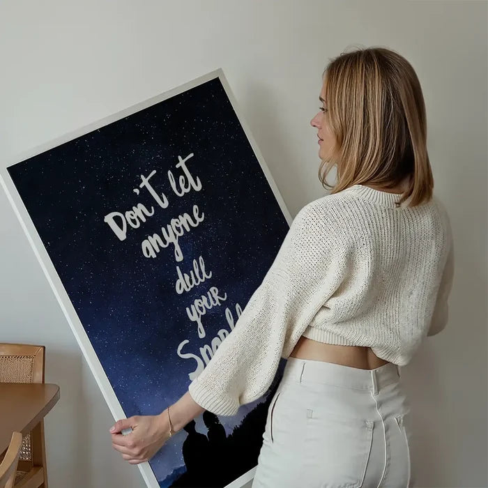 Woman holding a framed "Don't Let Anyone Dull Your Sparkle" print with a dark blue starry sky background.